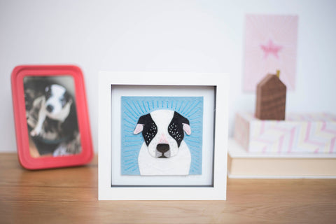 Portrait of dog. The dog is black and white and has embroidered detail on him. It's in a white frame and sits on a table.