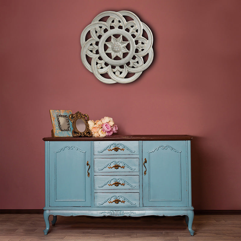 Mauve colored room with an antique wood accent cabinet with flowers and picture frames with a carved wood grey medallion hanging above. 