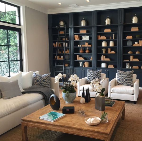 Living room next to a big window with a large square wood coffee table a cream colored couch, two cream arm chairs, throw pillows and a built in charcoal colored book case