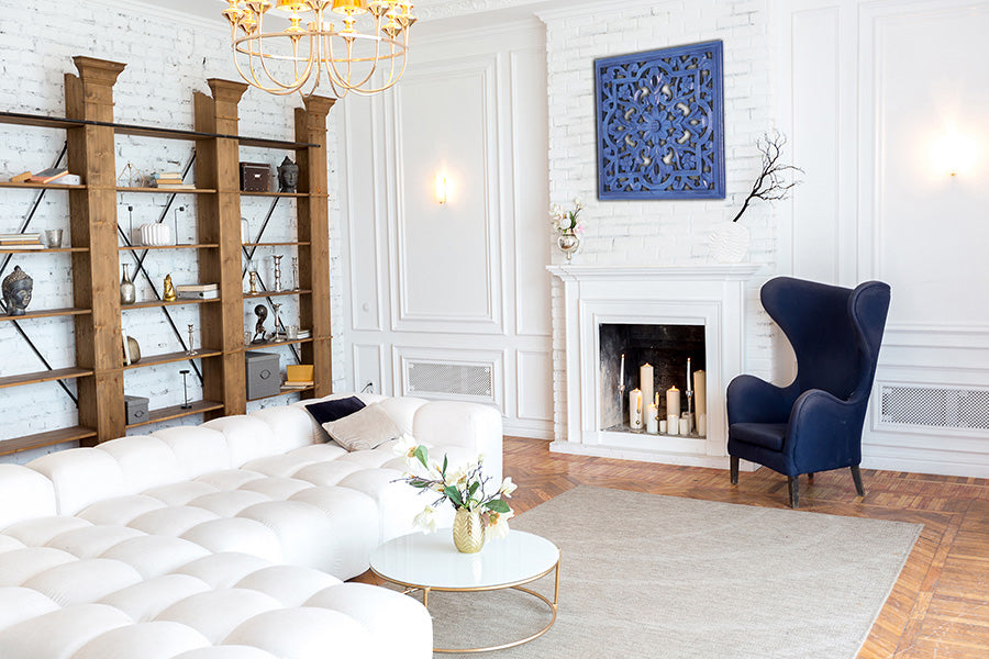 Room with white brick walls, light wood wall shelf, bright blue high back chair, blue carved wall medallion above mantel. 