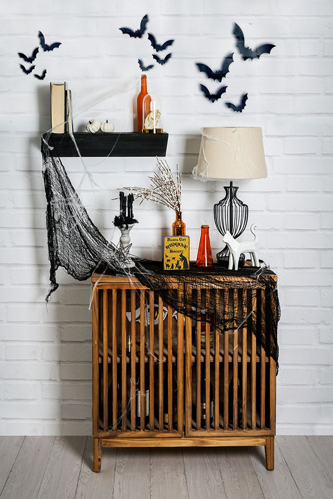 Minimal Halloween decor on a black wedge shelf and slated bar cabinet. Decorations include bats on the wall, black metal thick gauge wire lamp, spider webs, black webbing, ceramic cat figurine, and various bottles