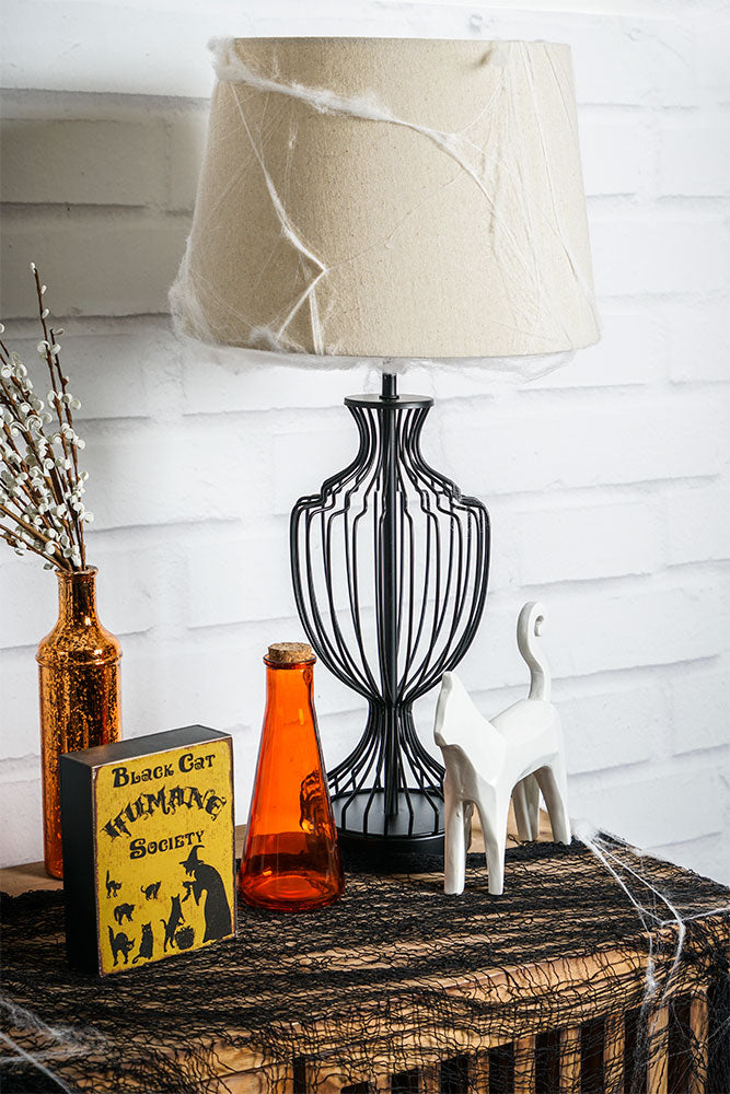 Black metal cage lamp on a wood slated cabinet surrounded by halloween decor including black webbing, spiderwebs on lampshade, orange bottles, plaque about black cats, and white ceramic cat statue