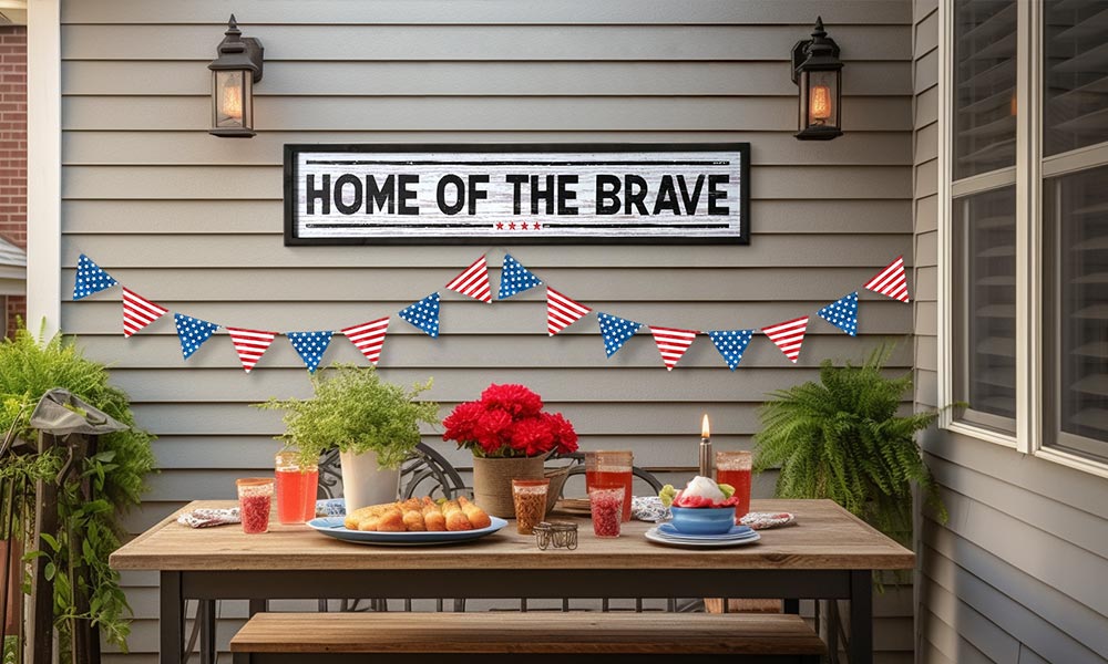 American Art Decor's Home of the Brave wood sign hanging on a patio wall by a table of food with an American flag banner.