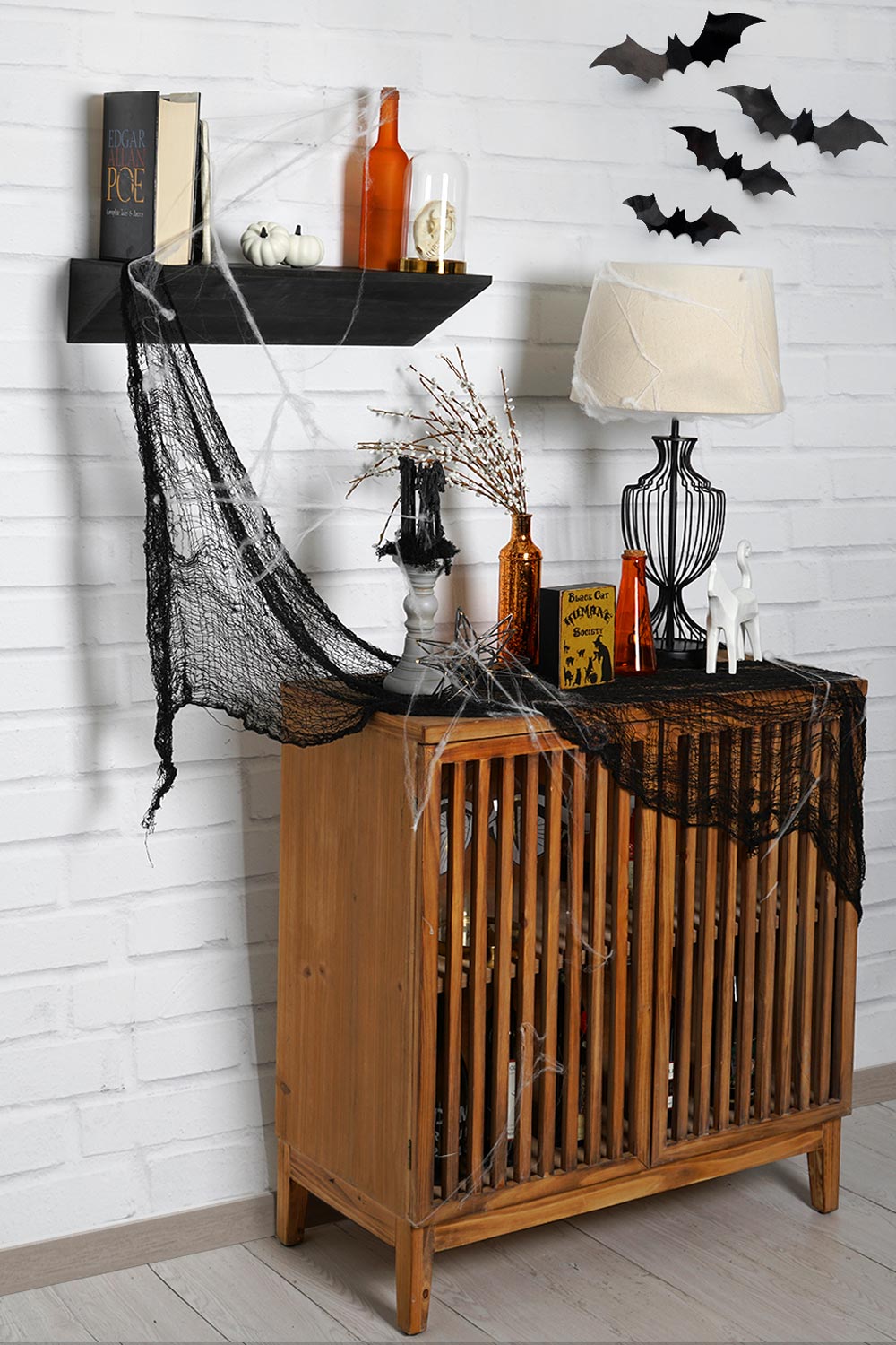 3/4 view of black wedge shelf with books and orange glass jars above a wood slated cabinet. Various Halloween decorations are on top of the cabinet and black bats are in upper right corner