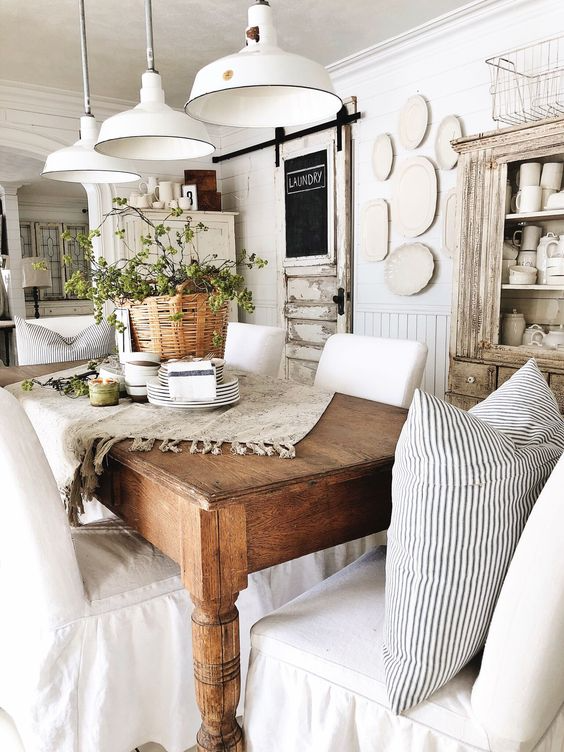 Dining room with an antique wood table chairs with cream chaircovers dishes piled on a table with a plant antique pendant lamps a vintage distressed white wood China cabinet and a wall of hanging plates