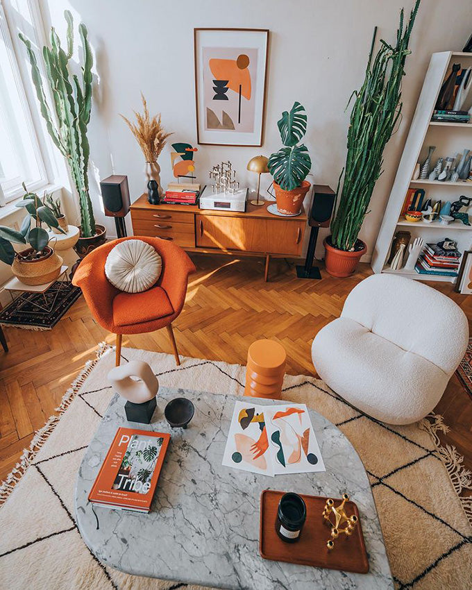 Living room with chevron hard wood floors, a marble coffee table on a cream area rug two vintage 1970s cushioned chairs in orange and white, a credenza with plants a lamp two small and two large cactus
