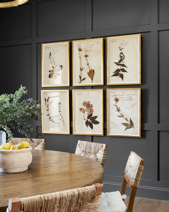 A round dining room table with rattan chairs, decorated with a fruit bowl and greenery. The paneled wall behind is painted in a dark green hue with six botanical images mounted to the wall.