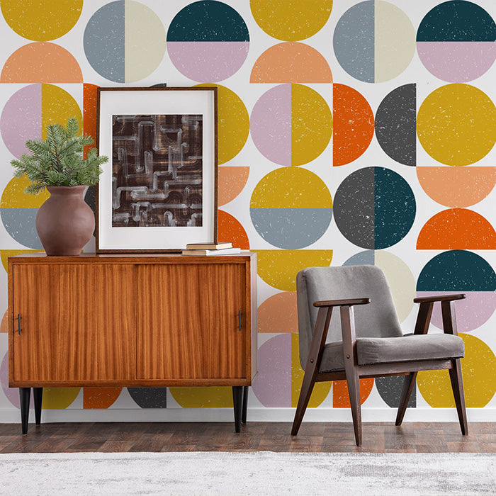A midcentury modern armchair in dark wood and grey upholstery is placed next to a mid-century modern credenza with a plant, and an abstract print with Geometric half circle wallpaper in mustard yellow, pink, navy, grey, cream and orange on a white background on the wall behind.