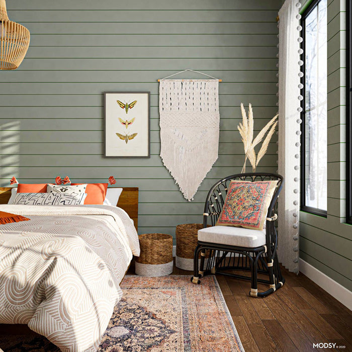 Sustainable bedroom decor, with large bedroom window, wicker rocking chair with floral throw pillow, distressed traditional area rug, macrame wall decor, sage green paneled wall, creme colored comforter and orange throw pillows.