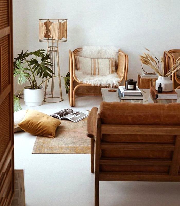 Room with a neutral colored area rug, glass table with books, an opened magazine and plant on the floor a rattan 1970s floor lamp, two rattan chairs and a wood chair with leather cushion