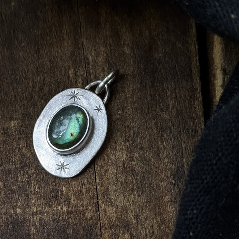 organic oval pendant with engraved star shapes and a bezel-set green labradorite in the center, on a dark wood background