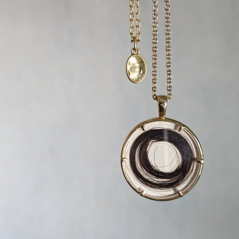 large circle pendant in yellow gold with a lock of hair inside, next to a small oval pendant with an eye made of strands of hair