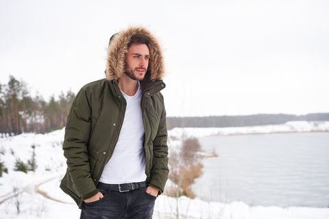 Handsome young man walking in the snow by a lake wearing a fur lined parka with hood