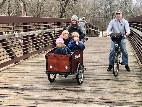 Mom cycling on a special needs trike carrying three kids across a wooden bridge with dad nearby on a two-wheeler. 
