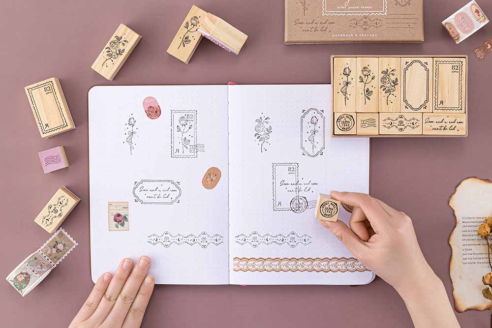 Tsuki ‘Vintage Rose’ Bullet Journal Stamp Set held in hands on open Tsuki ‘Vintage Rose’ Limited Edition Bullet Journal with Tsuki ‘Vintage Rose’ Washi Tapes with burnt paper and dried roses on mauve background