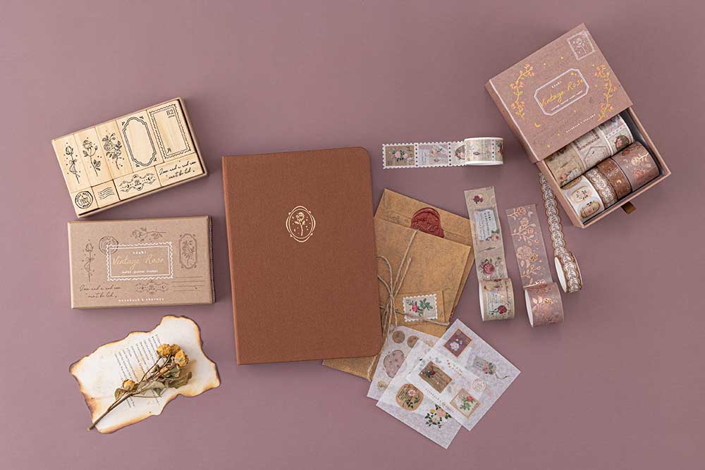 Tsuki ‘Vintage Rose’ Limited Edition Bullet Journal with Tsuki ‘Vintage Rose’ Washi Tape Set and Tsuki ‘Vintage Rose’ Bullet Journal Stamp Set with dried flowers and scrapbook letters and burnt paper on mauve background