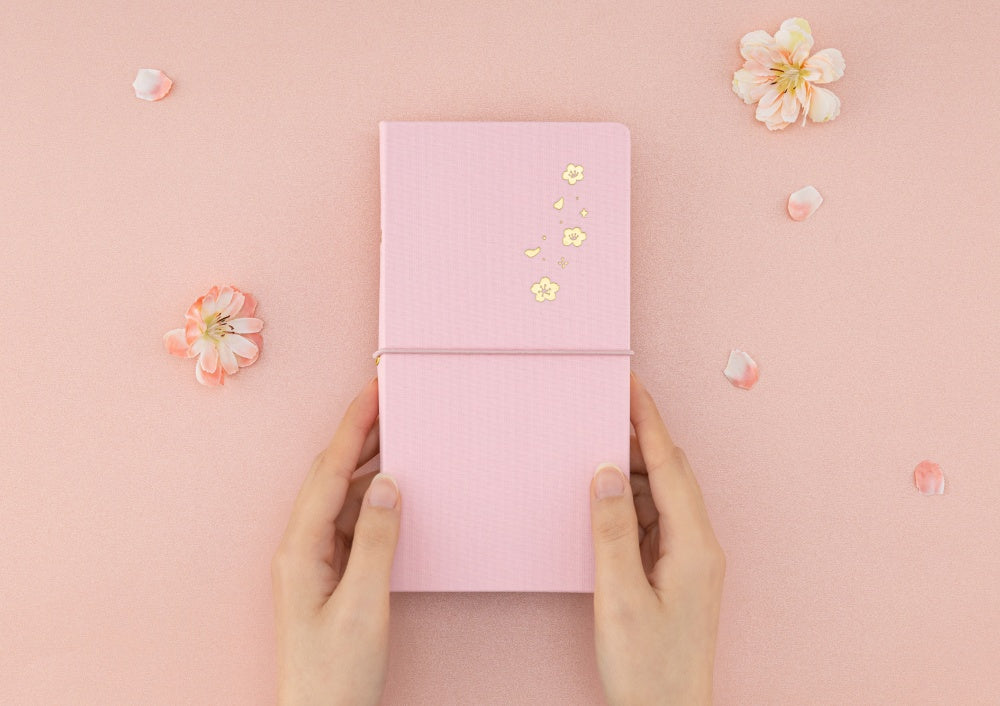 Hands holding travel notebook with pink linen cover and sakura gold foil detail