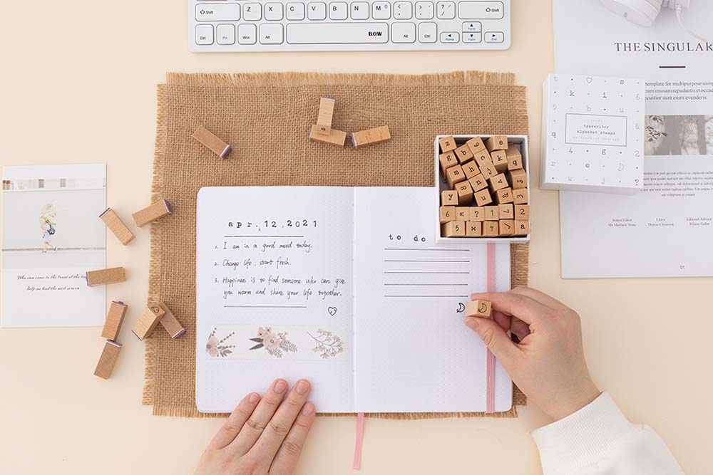 Tsuki Bullet Journal Typewriter Style Alphabet Stamps with eco-friendly gift box packaging and keyboard and headphones and paper being used on open bullet journal on hessian mat on beige background