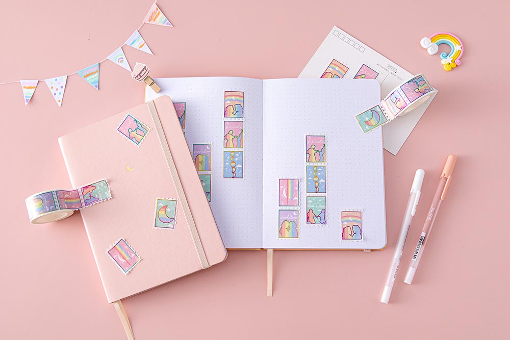 Tsuki Rainbow Pride Washi Tape on open notebook page with pink sakura Tsuki Pastel Edition Bullet Journal and postcard with rainbow and bunting and pens on light pink background