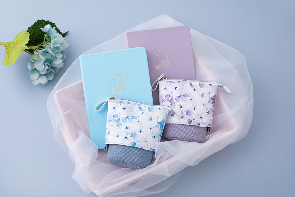 Tsuki Endless Summer Pop-Up pencil cases in petal blue and lilac bloom with Tsuki Endless Summer Bullet journals in basket with netting with blue hydrangea flowers on light blue background