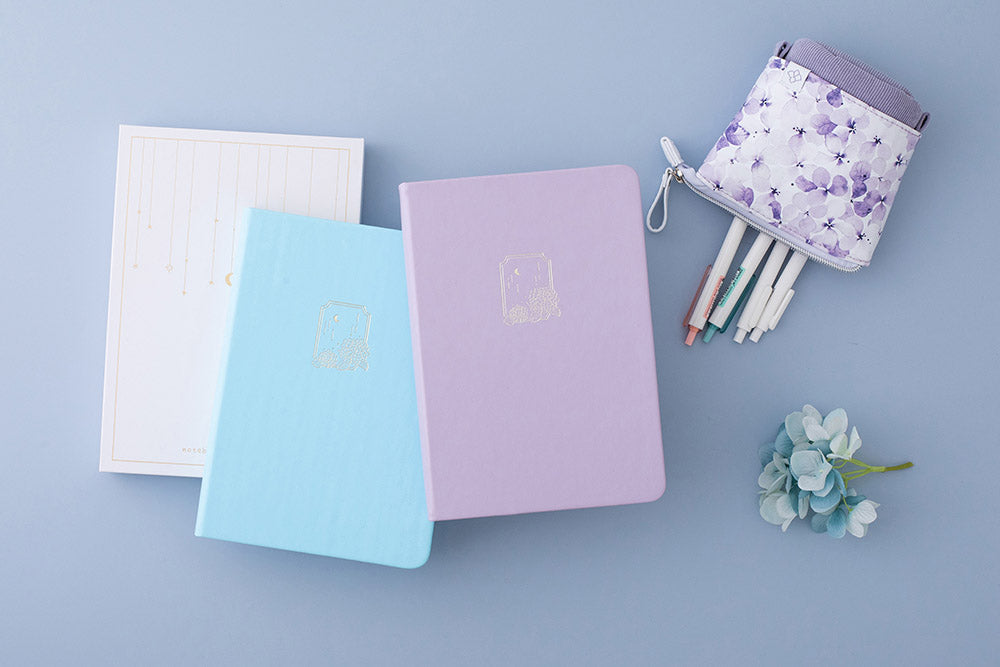 Tsuki Endless Summer Limited Edition Bullet Journals in Lilac Bloom and Petal Blue with eco-friendly gift box packaging and Tsuki Endless Summer Pop-Up Pencil case in Lilac Bloom and light blue hydrangea flowers on light blue background