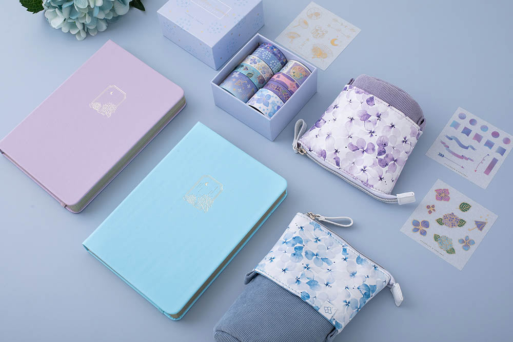 Tsuki Endless Summer Limited Edition Bullet Journals in Lilac Bloom and Petal Blue with Tsuki Endless Summer washi tape set and free sticker sheets with Tsuki Endless Summer Pop-Up Pencil cases with light blue hydrangea flowers at an angle on light blue background