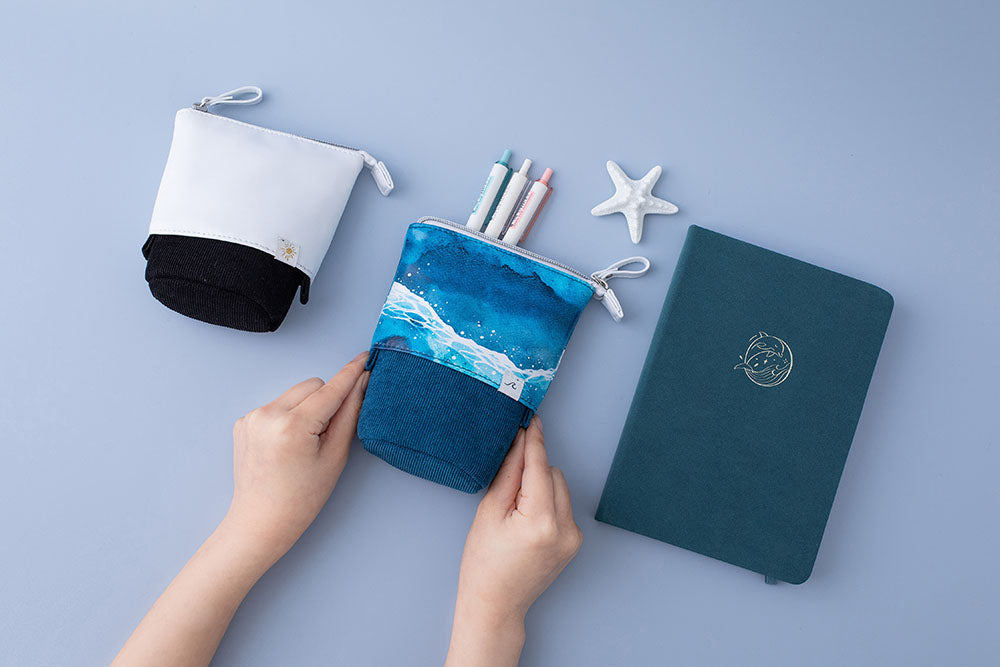 Tsuki Ocean Edition pop up standing pencil cases in Orca Black and Ocean Blue held in hands at an angle with sea green textured vegan leather Dolphin Days notebook with pens and starfish on blue background