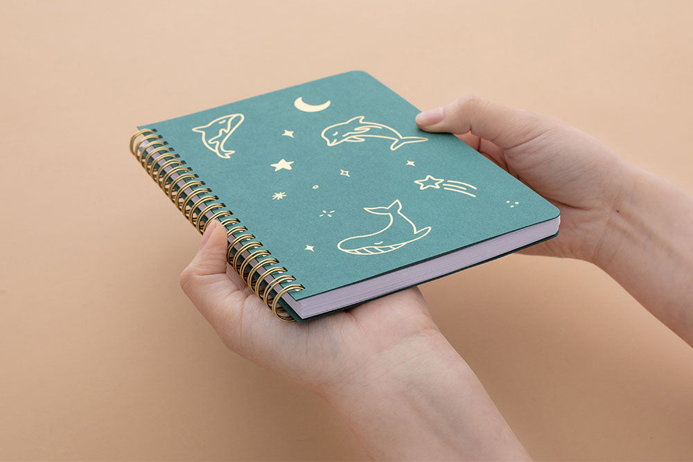 Tsuki Ocean Edition Ring Bound notebook in deep teal held in hands at an angle in peach background