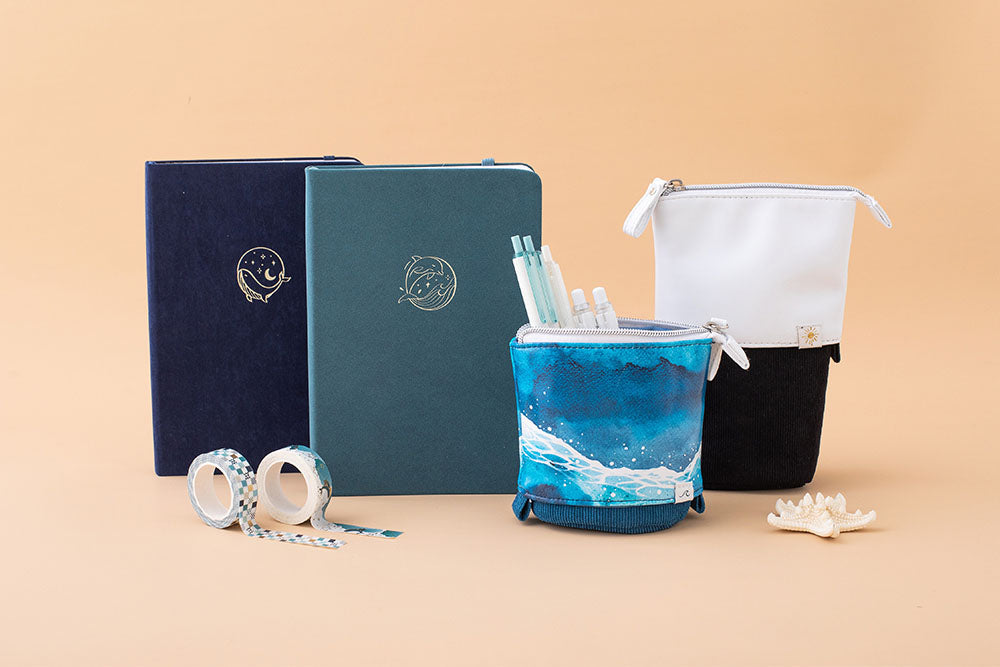 Tsuki Ocean Edition pop up standing pencil cases in Ocean Blue and Orca Black with Tsuki Ocean Washi Tape and sea green textured vegan leather Dolphin Days notebook and deep blue Gentle Giant luxury edition notebook with seashell in peach background