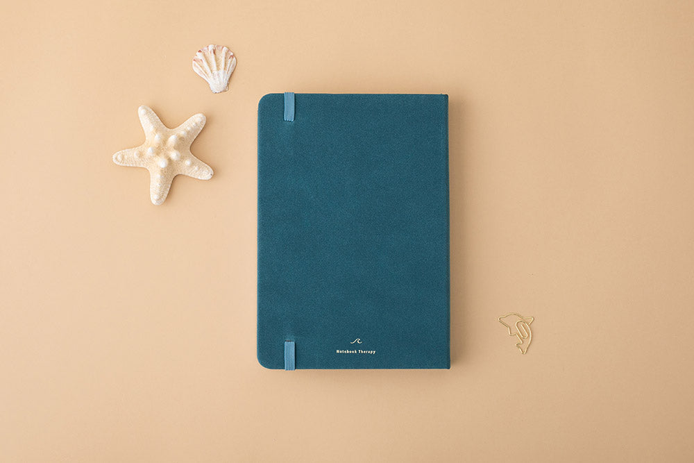 Focus on the back cover of Sea green textured leather Tsuki Dolphin Days bullet journal with starfish and seashell on peach background