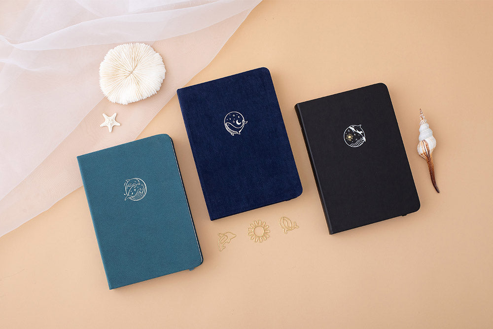 Tsuki sea green textured leather Dolphin Days notebook with Tsuki deep blue Gentle Giant notebook and Tsuki soft black Playful Orca notebook with seashells on peach background