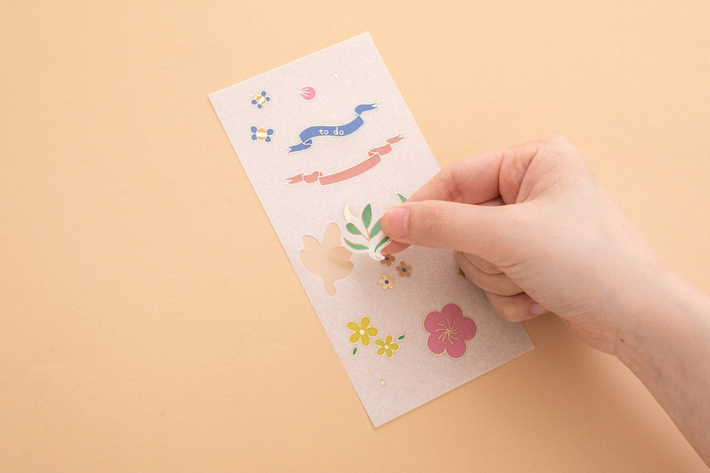 Hand peeling sticker off Tsuki Floral sticker sheet on peach background