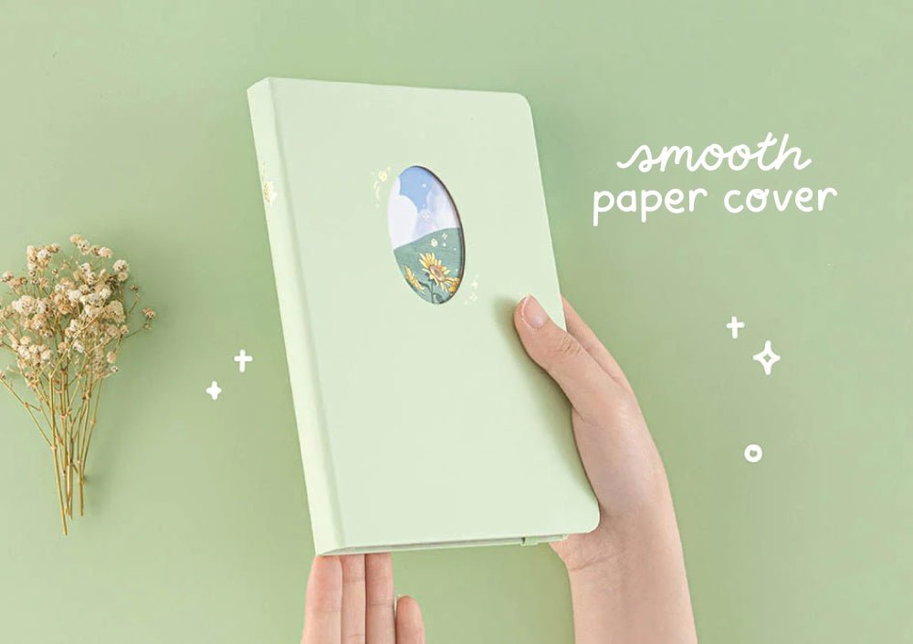 Hands holding Tsuki Four Seasons Summer Collectors Edition 2022 sage bullet journal notebook on sage green background with dried flower decoration on the left