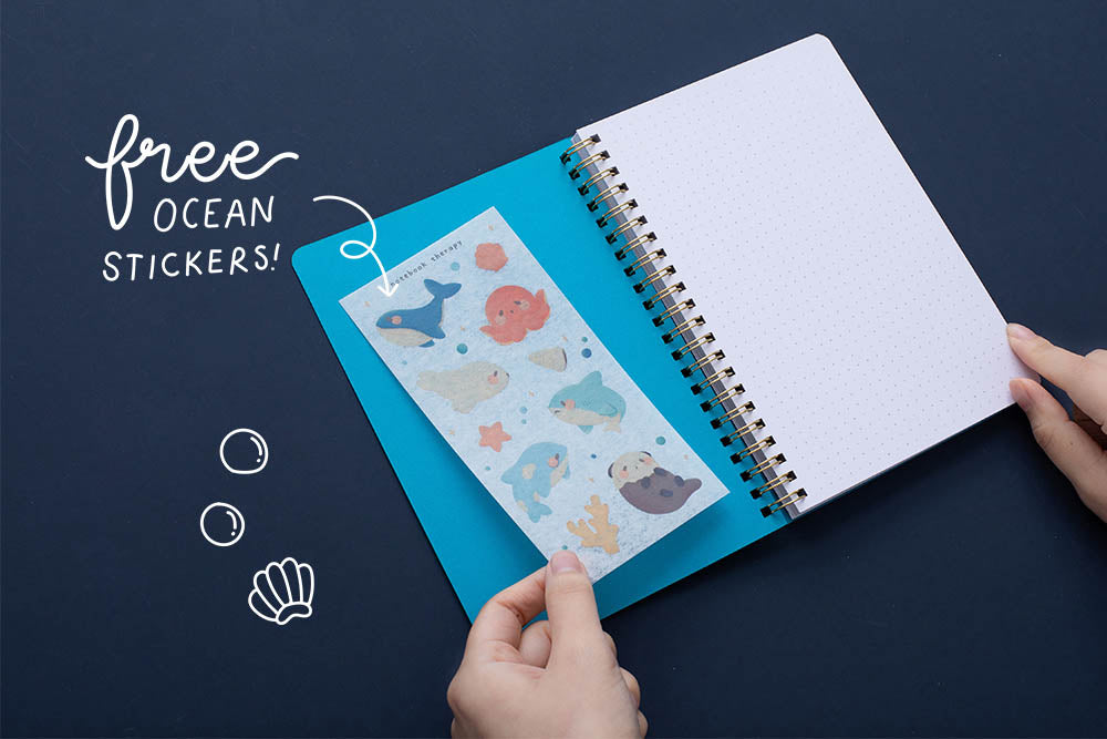 Tsuki Ocean Edition Ring Bound notebook in aqua blue open with free ocean stickers held in hands on dark blue background
