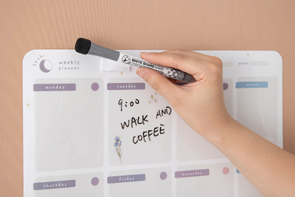 Close up of Tsuki Reusable Wall Planner with dry erase marker and free self-adhesive pen holder held in hand on smooth wallpaper surface