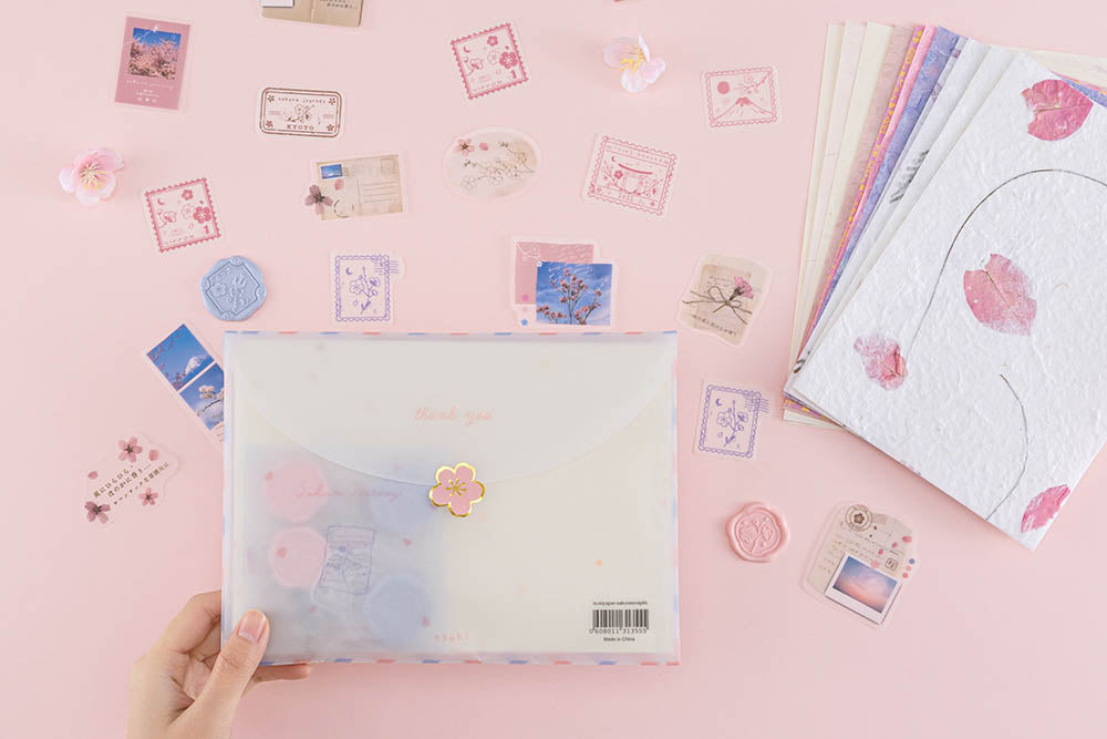 Tsuki ‘Sakura Journey’ Scrapbooking Set in envelope held in hands with cherry blossoms over pink background