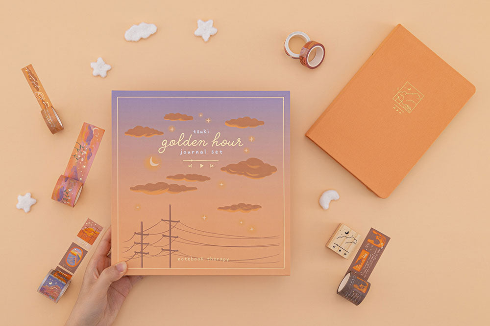 Hand holding the Tsuki Golden Hour Bullet Journal Box Set with the notebook and washi tapes and playlist stamp scattered in the orange background