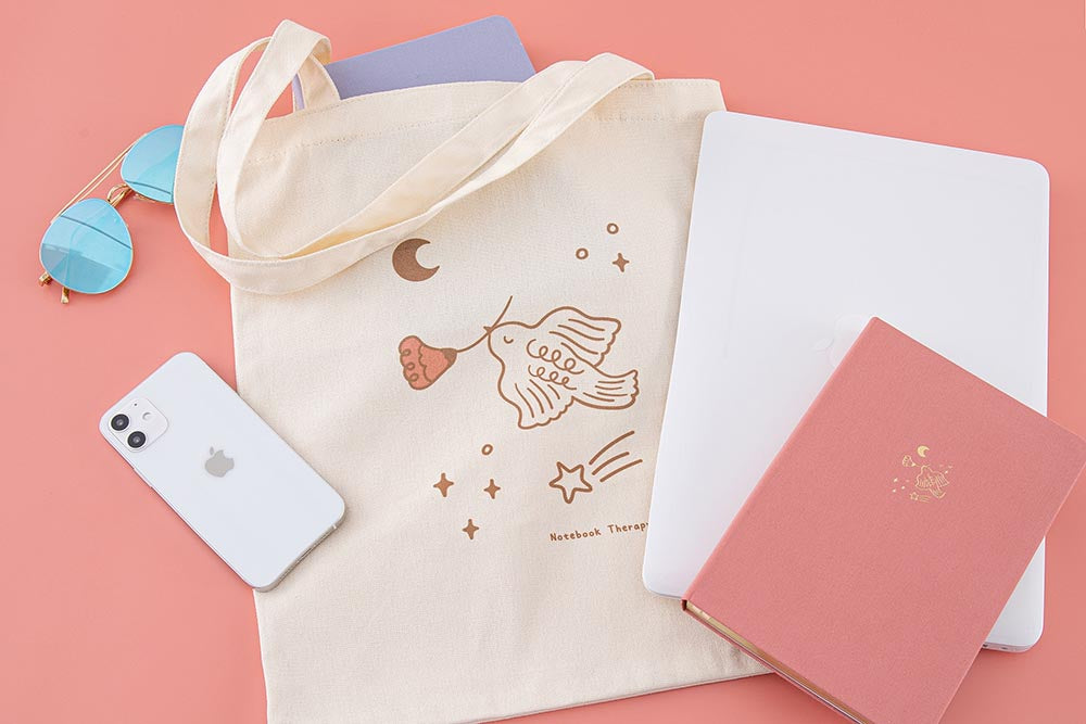 Tsuki ‘Moonflower’ Limited Edition Tote Bag with ‘Full Bloom’ Limited Edition Bullet Journal inside with ‘Suzume’ Limited Edition notebook and laptop and phone and sunglasses on coral pink background