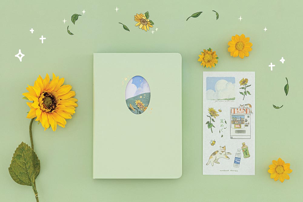 Flatlay of Tsuki Four Seasons Summer Collectors Edition 2022 sage bullet journal notebook next to the free sticker sheet and sunflower decorations on sage green background