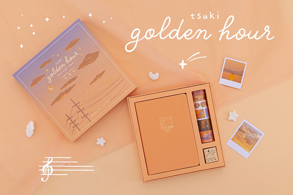 Flatlay of Tsuki Golden Hour bullet journal box set, box is opened revealing the contents which include a notebook, 6x  washi tapes and a wooden rubber stamp  on orange background with sunset polaroids scattered in the background