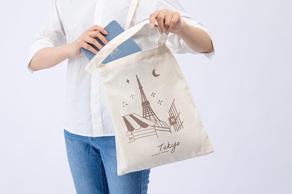 A figure carrying a Tokyo tote bag and putting a blue notebook inside the bag