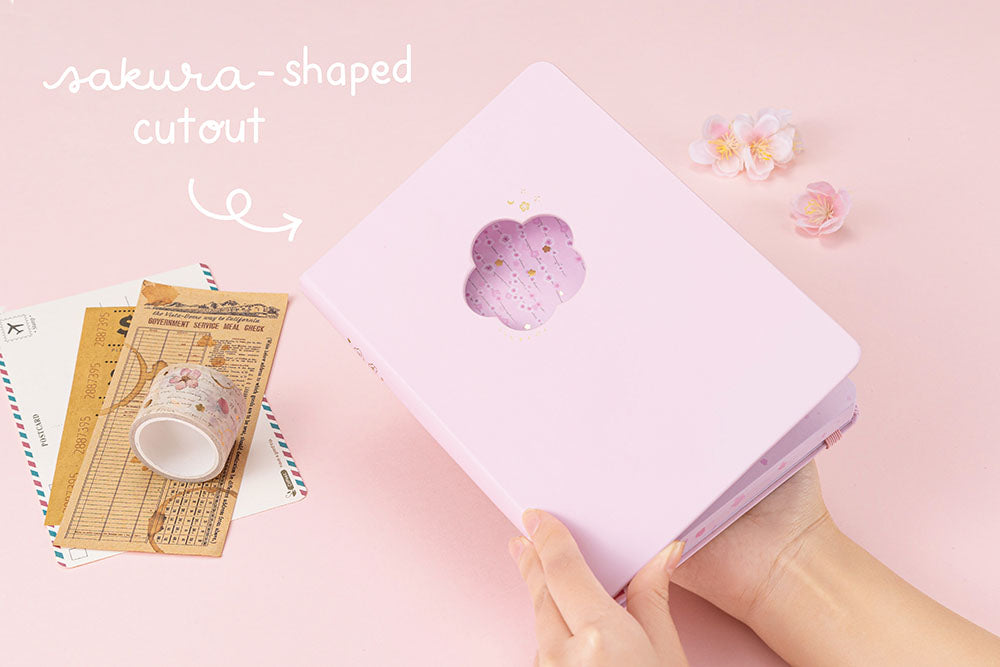 Tsuki Four Seasons: Spring Collector’s Edition 2022 Bullet Journal with sakura shaped cut out held in hands with Tsuki ‘Sakura Journey’ Vintage Journal Washi Tape Set with travel notes and postcard and sakura blooms on light pink background