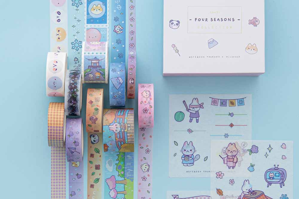 Close up of Tsuki ‘Four Seasons’ Washi Tape Set by Notebook Therapy x Milkkoyo rolled out with three free stickers sheets on light blue background
