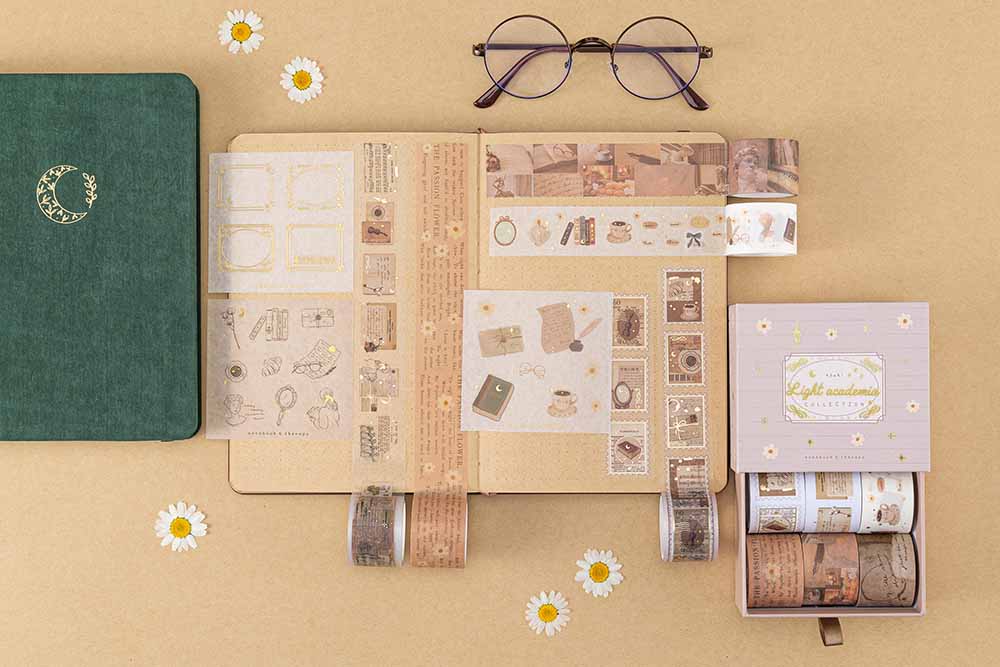 Tsuki Light Academia Washi Tape set on kraft paper bujo with the open drawer box next to it and the green velvet bullet journal and a pair of glasses and some pressed daisies scattered around on brown background