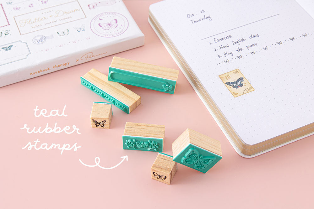 Close up of Tsuki ‘Flutter + Dream’ Bullet Journal Stamp Set by Notebook Therapy x Pelinkan with teal rubber stamps with open Tsuki Cloud White ‘Flutter + Dream’ Limited Edition Bullet Journal by Notebook Therapy x Pelinkan on pastel pink background