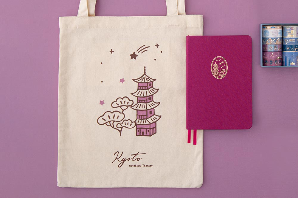 A flatlay of a Kyoto tote bag and a fuchsia pink notebook with a temple design