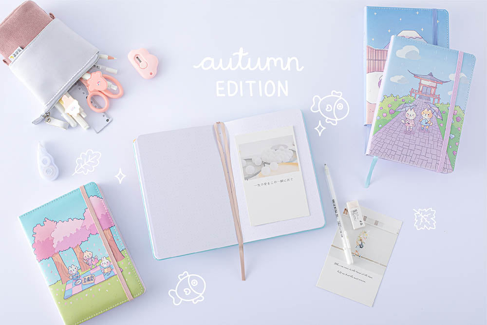 Tsuki ‘Four Seasons: Autumn Edition’ Bullet Journal made with milkkoyo held in hand with Tsuki ‘Four Seasons: Summer Edition’ notebook and Tsuki ‘Four Seasons: Spring Edition’ bujo and Tsuki ‘Four Seasons: Winter Edition’ bullet journal with Tsuki Pop-Up Pencil case and assorted stationery supplies on lilac background