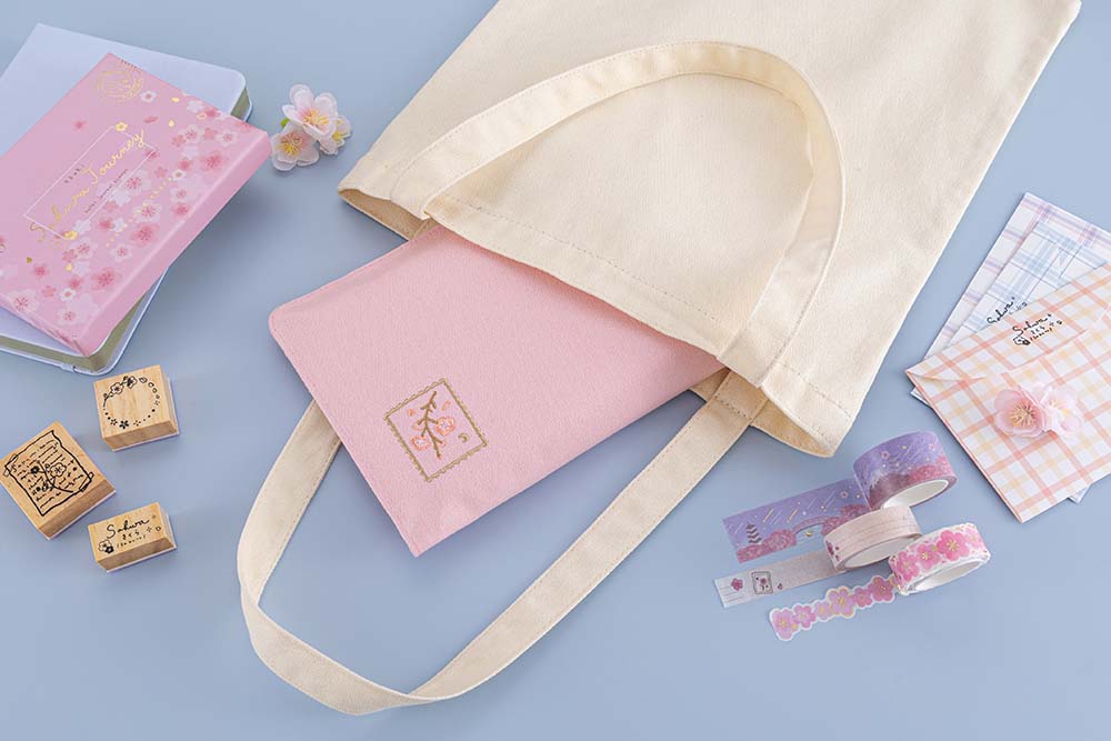 Tsuki ‘Sakura Journey’ Notebook Pouch inside canvas tote bag with Tsuki ‘Sakura Journey’ Bullet Journal Stamp Set and Tsuki ‘Sakura Journey’ Limited Edition Bullet Journal and Tsuki ‘Sakura Journey’ Washi Tapes with gingham envelopes and cherry blossoms on light blue background