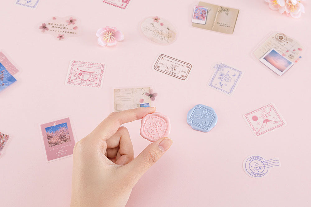 Wax seal sticker from Tsuki ‘Sakura Journey’ Scrapbooking Set held in hand over sticker flakes on pink background