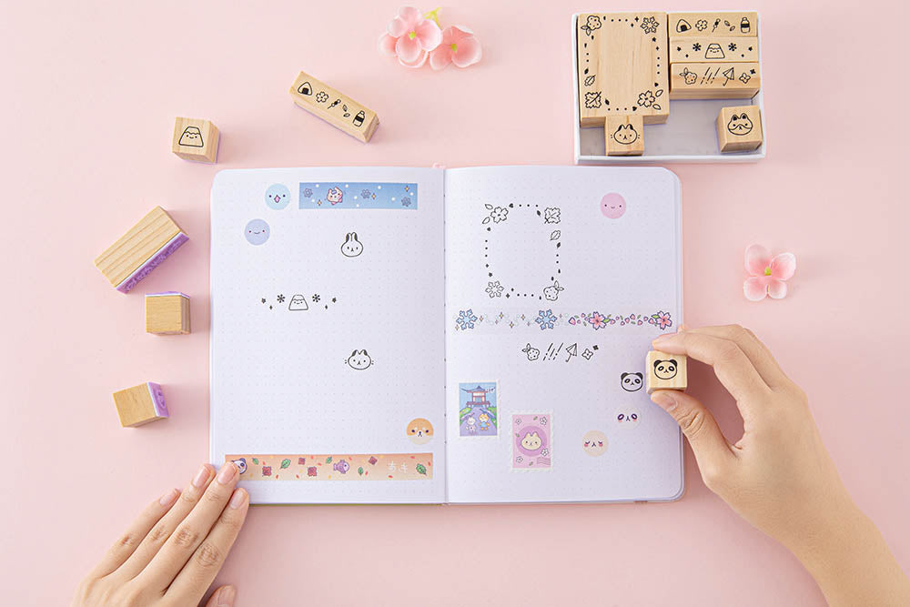 Tsuki ‘Four Seasons’ Bullet Journal Stamp Set by Notebook Therapy x Milkkoyo held in hands over open bullet journal page spread with Tsuki ‘Four Seasons’ Washi Tapes with pink flowers on petal pink background